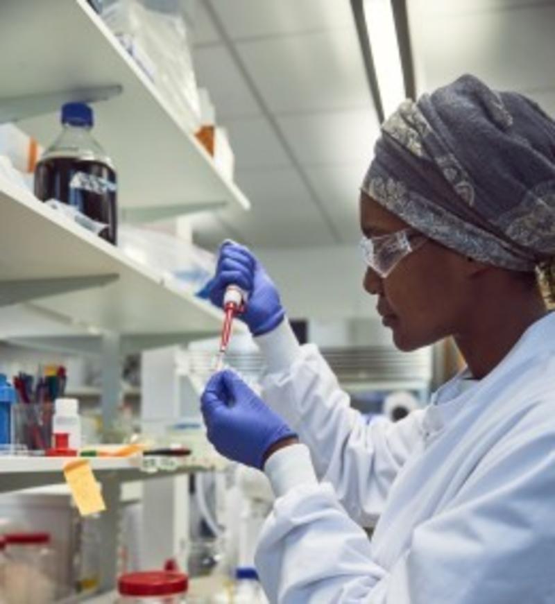 A woman of African heritage in full lab clothing looking at a sample in her lab