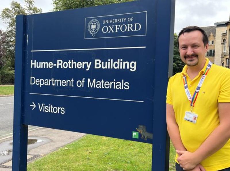 Dr Greg Mazur smiling and leaning against the main directional sign sign posting the the Department of Materials' building in Parks Road