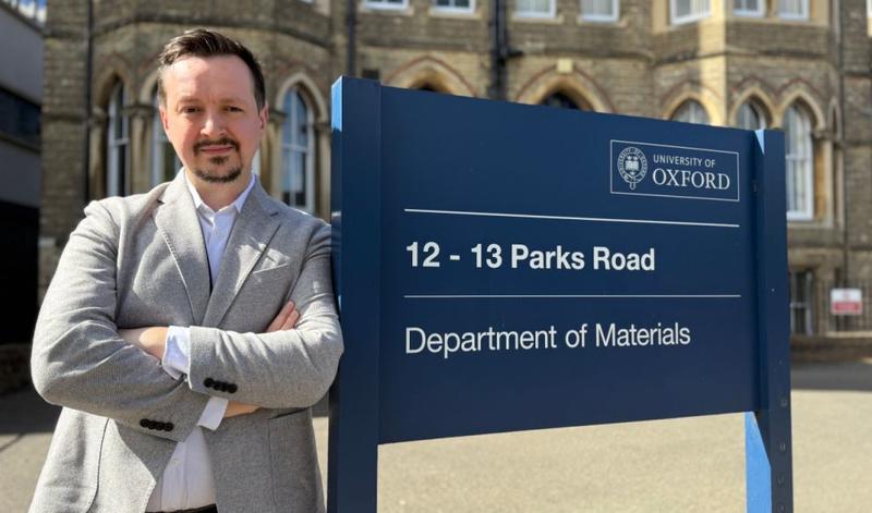 Dr Greg Mazur looking serious and leaning again the departmental sign outside 12 13 Parks Road