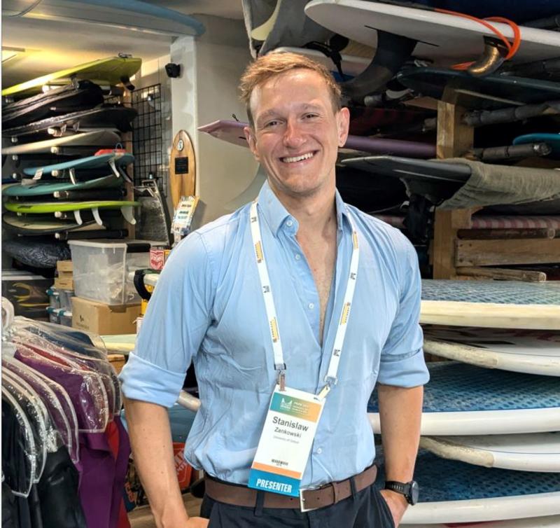 Half body shot of Dr Zankowski smiling, wearing his conference lanyard and standing in a room containing surf boards and related clothing and accessories