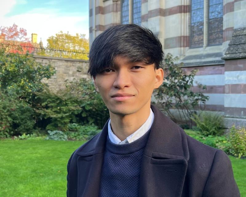 Head and shoulders of Hiiro Moriyama, standing in a walled Oxford garden, with a Victorian gothic building behind (possibly a part of Keble College)