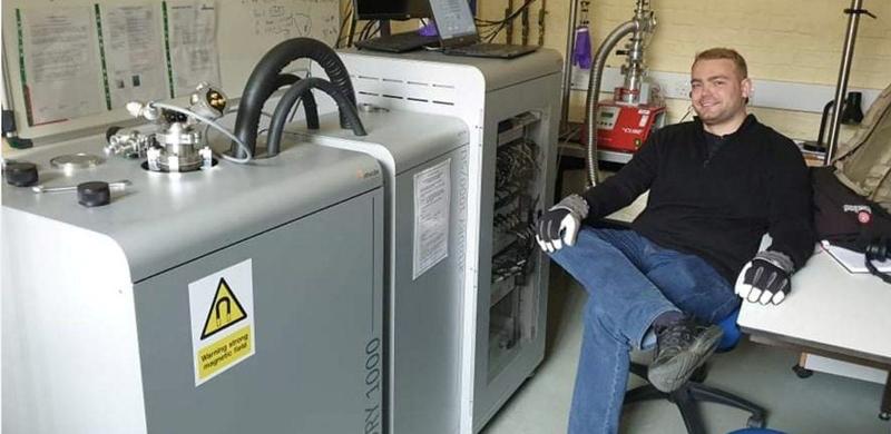 Dr Petr Zaguar sitting in a lab and smiling.  There are some large items of equipment by him and he is wearing protective gloves