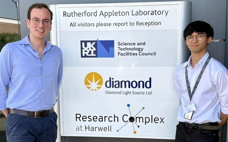 Stefano and Hiiro standing either side of the ral signage for UKRI Diamond and Harwell research complex