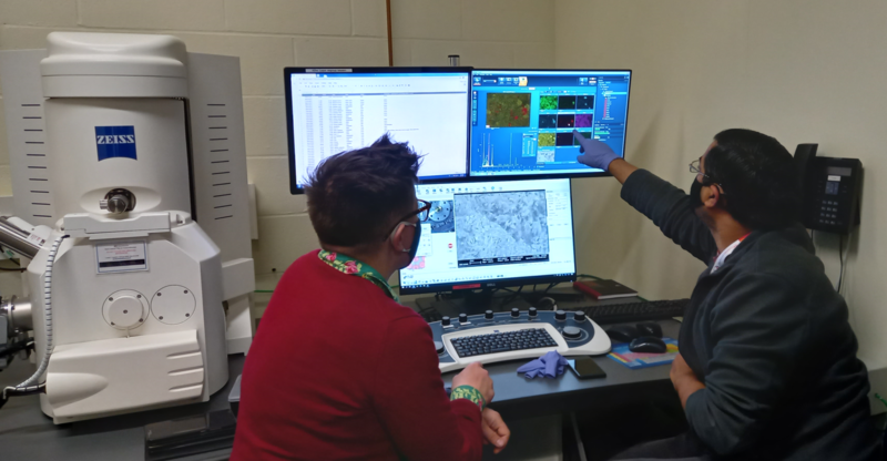 The backs of Dr Katrin Wilhelm and Dr Phani Karamched as they face two monitors full of SEM sample data. There is a keyboard on the desk and a Zeiss machine to one side