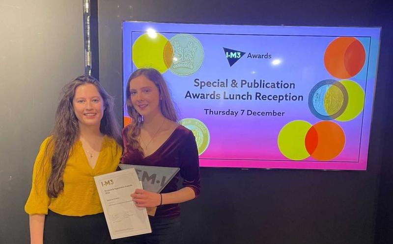 Dr Leanne Jones and Rosanna Roskilly standing in front of an LED displaying branding the prizes.  Rosanna is holding her award and prize certificate.